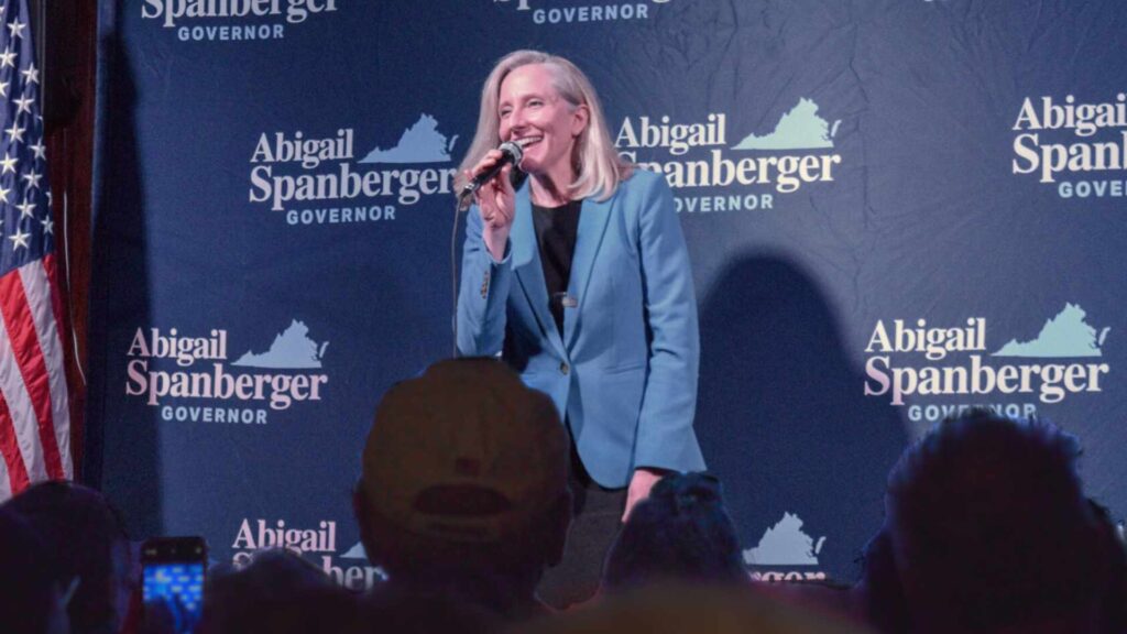 Abigail Spanberger speaking with microphone in front of step turn with her 2025 gubernatorial campaign logo. She is wearing a light blue blazer. An American flag is next to her.